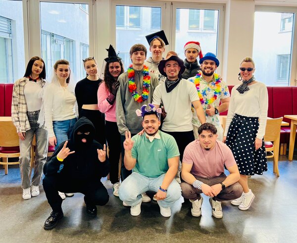 Eine größere Gruppe von Menschen in einem Raum mit großen Fenstern, die in verschiedenen Kostümen und Accessoires verkleidet sind. Manche tragen Hüte, Blumenketten oder Masken, während andere kreative Outfits wie ein schwarz vermummtes Gesicht oder eine Retro-Kleidung gewählt haben. Alle lächeln und posieren fröhlich für das Gruppenfoto.