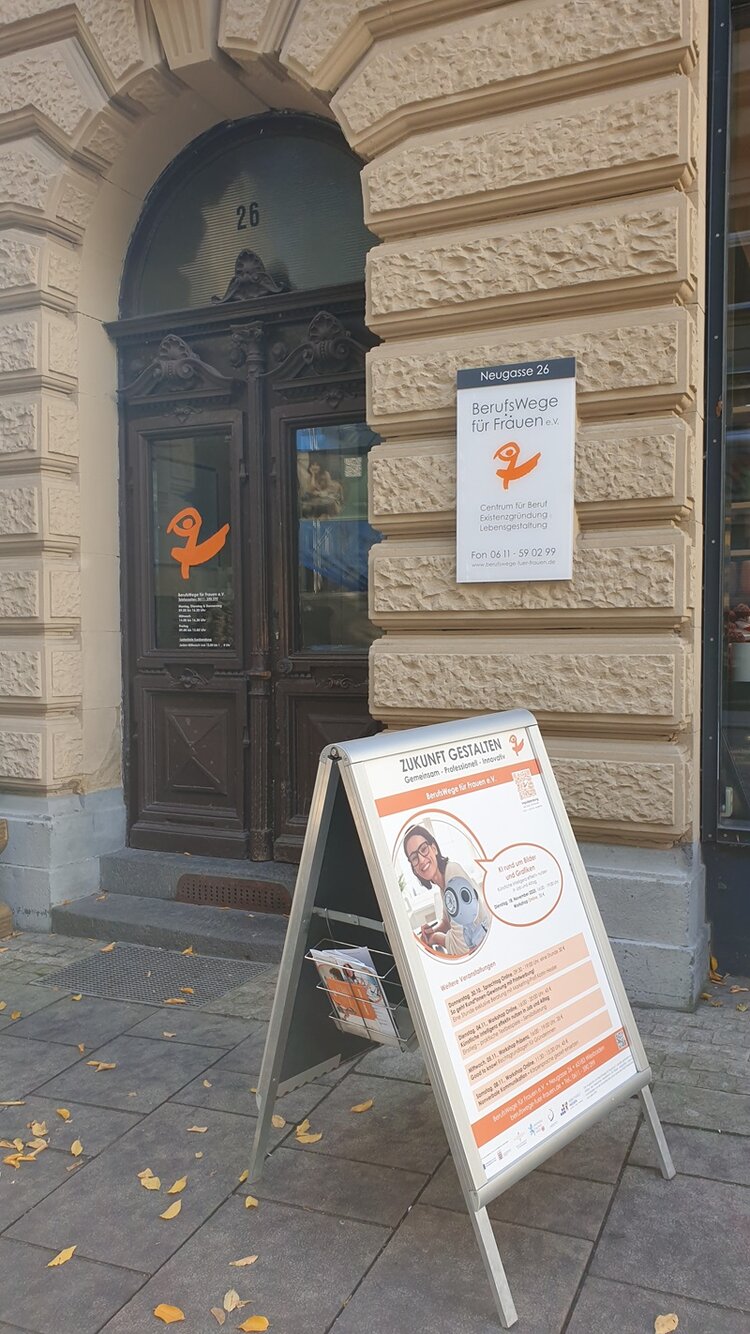 Vor einem historischen, sandsteinfarbenen Gebäude mit der Hausnummer 26 steht ein dunkelbraunes, zweiflügeliges Holztor mit verzierten Rahmen. Rechts neben dem Tor ist ein weißes Schild mit dem orangefarbenen Logo und der Aufschrift 'BerufsWege für Frauen e.V.' angebracht. Vor dem Eingang steht ein aufklappbares, weißes Werbeaufsteller-Schild mit orangefarbenen und schwarzen Texten sowie einem Bild, das auf eine Veranstaltung oder ein Angebot hinweist. Der Gehweg ist mit herabgefallenen gelben Blättern bedeckt, was auf Herbst hinweist. Die Szene zeigt den Eingangsbereich der Euro-Schulen Wiesbaden im Rahmen einer Exkursion zur BerufsWege für Frauen e.V.