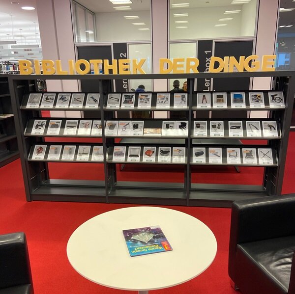 Blick auf die Bibliothel der Dinge, ein großes Regal mit Infobroschüren, roter Teppichboden, ein Tisch und zwei Ledersessel.