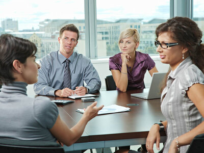 Vier Personen sitzen in einem modernen Besprechungsraum mit großen Fenstern und einer Stadtansicht im Hintergrund. Zwei Männer und zwei Frauen nehmen an einer Besprechung teil. Die Frau links, mit einem grauen Pullover, erklärt etwas und gestikuliert mit der Hand. Der Mann in der Mitte, in einem grauen Hemd, hört aufmerksam zu und hat einen Stift in der Hand. Die Frau rechts, mit Brille und einem grauen Oberteil, sitzt mit verschränkten Armen und hört ebenfalls zu. Die Frau in der Mitte, mit hellblonden Haaren und einem lila Oberteil, stützt ihr Kinn auf die Hand und wirkt nachdenklich. Auf dem Tisch liegen Unterlagen, ein Laptop und ein Handy. Die Atmosphäre wirkt professionell und konzentriert.
