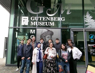 Vor dem Eingang des Gutenberg-Museums stehen die Teilnehmenden der Euro-Schulen Mainz und lächeln für ein Gruppenfoto. Die Glasfassade zeigt großflächig den Schriftzug 'GUTENBERG MUSEUM' sowie ein Porträt von Johannes Gutenberg als schwarz-weiße Linienzeichnung. Die Teilnehmenden tragen unterschiedliche Freizeitkleidung und blicken in Richtung Kamera. Im Hintergrund sind weitere Schriftzüge und das Museumslogo sichtbar.