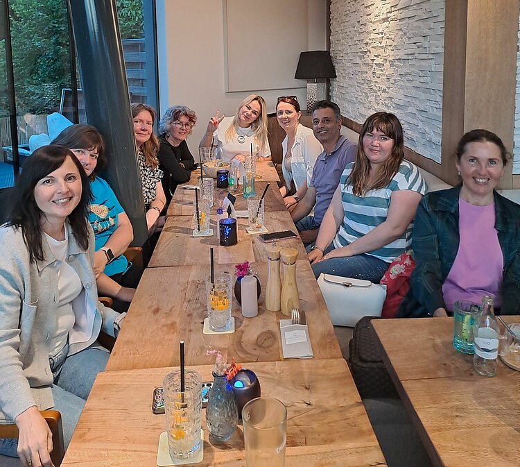 Eine Gruppe von neun Personen sitzt gemeinsam an einem langen Holztisch in einem Restaurant. Auf dem Tisch stehen Getränke und Gläser. Alle lächeln in die Kamera, die Stimmung wirkt entspannt und freundlich.