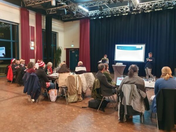 In einem Veranstaltungssaal sitzen mehrere Personen an runden Tischen und schauen nach vorne zu einer Leinwand, auf der eine Präsentation läuft. Zwei Vortragende stehen nebeneinander, während im Hintergrund ein dunkler Bühnenvorhang und Scheinwerfer zu sehen sind.