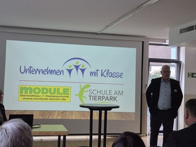 Ein Referent steht neben einer Projektionsfläche mit der Präsentation „Unternehmen mit Klasse“ und Logos verschiedener Partner.
