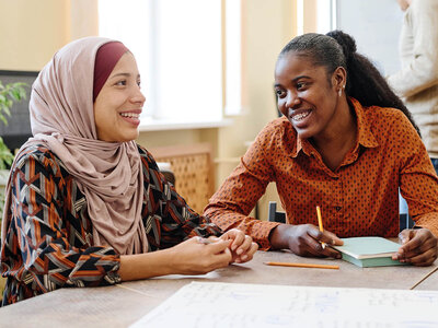 Zwei Frauen sitzen an einem Tisch und arbeiten zusammen. Die Frau links trägt ein Kopftuch und ein gemustertes Oberteil, während die Frau rechts ein orangefarbenes, gepunktetes Hemd trägt und in ein Notizbuch schreibt. Beide lächeln und wirken konzentriert auf ihre Aufgabe.
