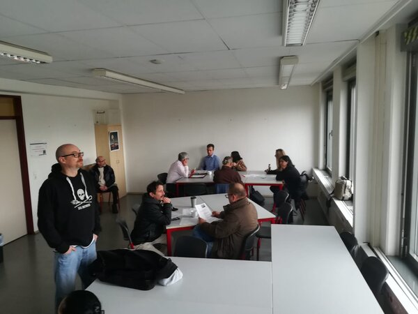 Überblick über einen Raum mit mehreren Gesprächsgruppen an Tischen; links steht eine Person, rechts lange Fensterreihe.