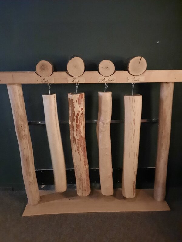 Fünf senkrecht aufgehängte Holzstämme mit naturbelassener Rinde und sichtbaren Jahresringen hängen an Haken von einer waagerechten Holzleiste. Über jedem Stamm ist auf der Leiste handschriftlich ein Wort notiert. Die Stämme unterscheiden sich in Maserung und Rindenanteil. Der Hintergrund ist dunkel, der Boden grau.