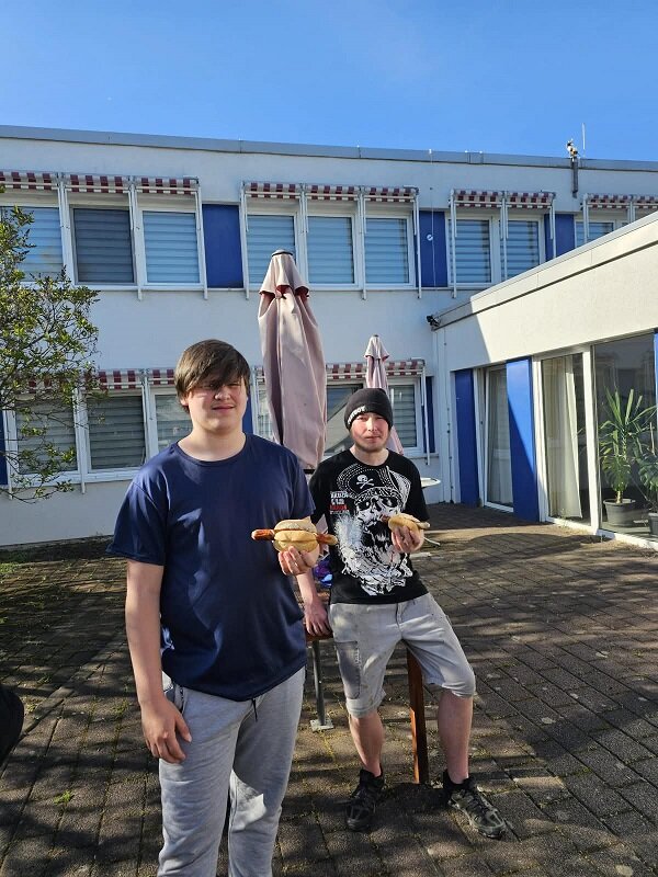 Zwei Personen posieren lächelnd vor einem Gebäude mit blauen Fensterrollläden. Beide halten ein Brötchen mit einer Bratwurst in der Hand. Im Hintergrund sind ein geschlossener Sonnenschirm und Pflanzen zu sehen.
