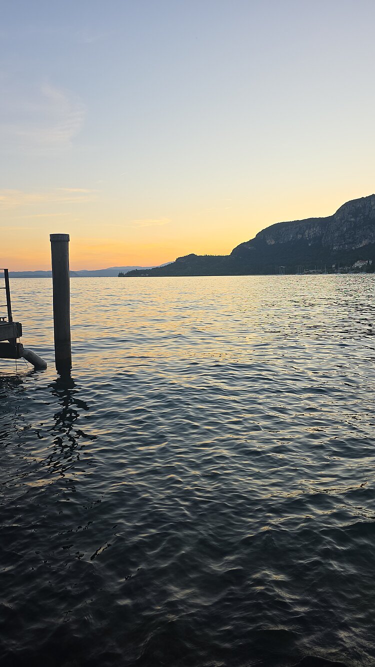 Zwischen Cocktail- und Regenschirm Blick auf einen Bergsee im Sonnenuntergang. An der Seite des Bildes lässt sich ein Steg erkennen. Die untergehende Sonne spiegelt sich im Wasser.