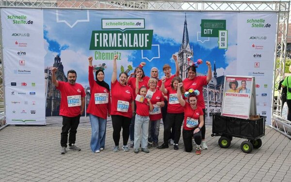 Gruppenbild eines jubelnden Teams in roten Euro-Schulen-Laufshirts vor einer Wand mit der Aufschrift "Firmenlauf Chemnitz". Neben der Gruppe steht außerdem ein kleiner Bollerwagen mit einem Poster zum Thema "Deutsch lernen".