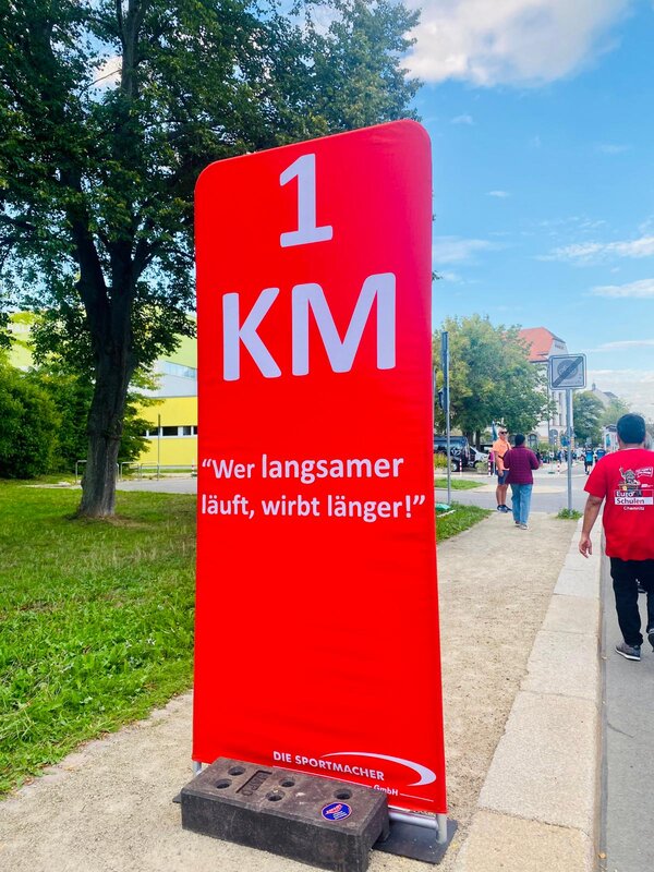 An einer Straße steht ein roter Aufsteller mit der Aufschrift: 1 KM "Wer langsamer läuft, wirbt länger!"