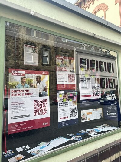 Schaufenster mit mehreren Plakaten und Flyern, die dicht nebeneinander an der Innenseite der Scheibe angebracht sind. Die Plakate zeigen überwiegend weiße Hintergründe mit roten und blauen Akzenten sowie großflächigen QR-Codes. Auf einigen Plakaten sind Porträtfotos von Menschen zu sehen. Die Texte auf den Plakaten informieren über Bildungsangebote und Ausbildungen, unter anderem mit dem Hinweis 'DEINE ZUKUNFT STARTET JETZT!'. Im linken Bereich hängt eine rote Tragetasche mit weißem Aufdruck. Die Spiegelung der gegenüberliegenden Häuserfront ist in der Scheibe sichtbar.
