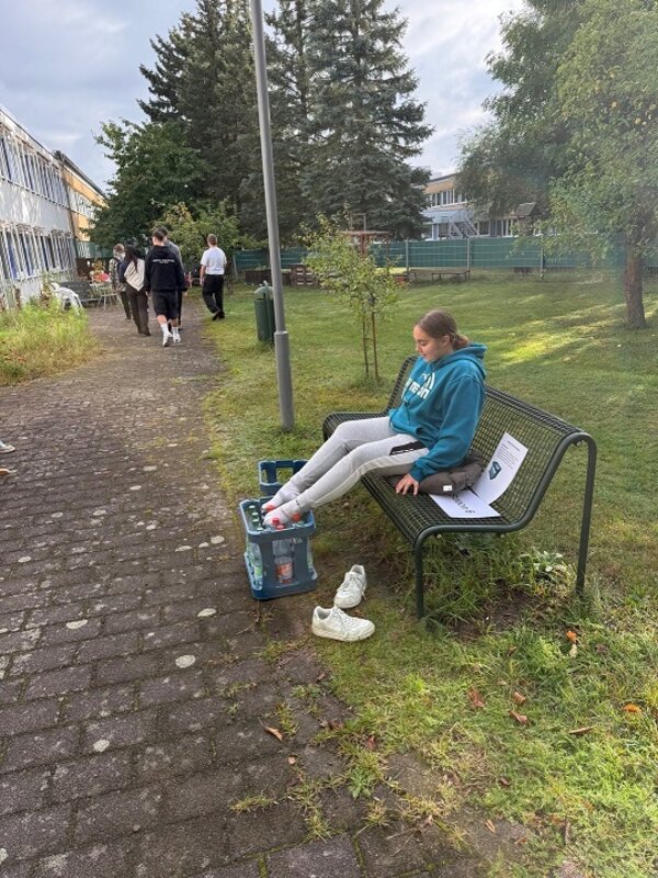 Eine Auszubildendemit türkisfarbener Kapuzenjacke und grauer Jogginghose sitzt auf einer Metallbank im Grünen. Sie hat die Beine auf zwei Getränkekisten gelegt und trägt keine Schuhe. Neben der Bank stehen weiße Turnschuhe. Auf der Bank liegt ein Blatt Papier. Im Hintergrund gehen mehrere Personen auf einem gepflasterten Weg an Gebäuden und Bäumen vorbei.