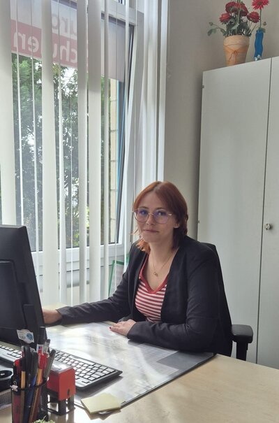Die ehemalige Auszubildende und nun neue Kollegin Lea sitzt vor dem Computer am Schreibtisch, trägt eine Brille, ein weiß-rot gestreiftes Oberteil und einen Blazer darüber und lächelt in die Kamera. 