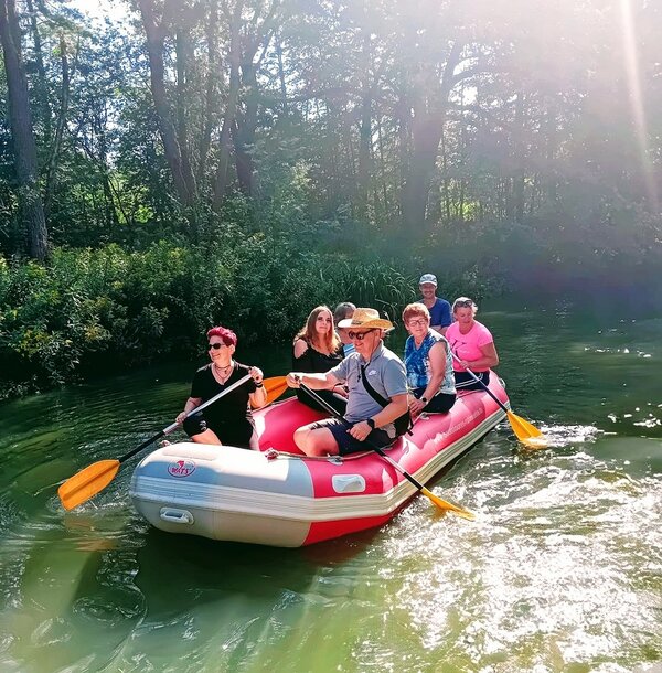 In einem roten Schlauchboot fahren sieben Personen den Fluss entlang. Die beiden Männer tragen einen Cowboyhut und eine Baseballkappe. Vier Personen halten ein Ruder in der Hand. Das Ufer ist mit Schilfgras, Büschen und Bäumen bewachsen.