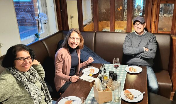 Ein männlicher und zwei weibliche Mitarbeiter*innen der Euro-Schulen bzw. Euro Akademie, die zusammen an einem Tisch in einem Restaurant sitzen und etwas essen und in die Kamera lächeln.