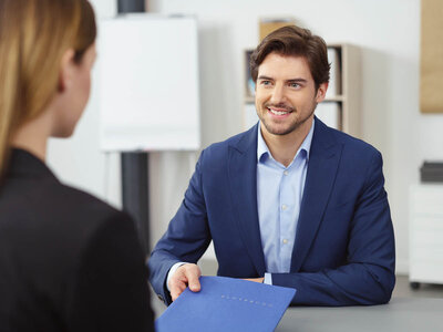 Ein Mann in einem dunkelblauen Anzug und einem hellblauen Hemd sitzt an einem Tisch und lächelt eine Frau an, die ihm gegenübersitzt. Er hält eine blaue Mappe in der Hand, was auf ein formelles oder berufliches Treffen hinweist.