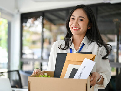 Eine Frau mit langen, dunklen Haaren und einem weißen Hemd steht in einem Büro oder einer Arbeitsumgebung. Sie trägt ein Namensschild um den Hals und hält einen Karton mit verschiedenen Gegenständen, darunter ein schwarzes Notizbuch, eine braune Mappe und einige Papiere. Sie lächelt und scheint zufrieden oder glücklich zu sein. Der Hintergrund ist unscharf, zeigt aber einen modernen Innenraum mit großen Fenstern und Büromöbeln.