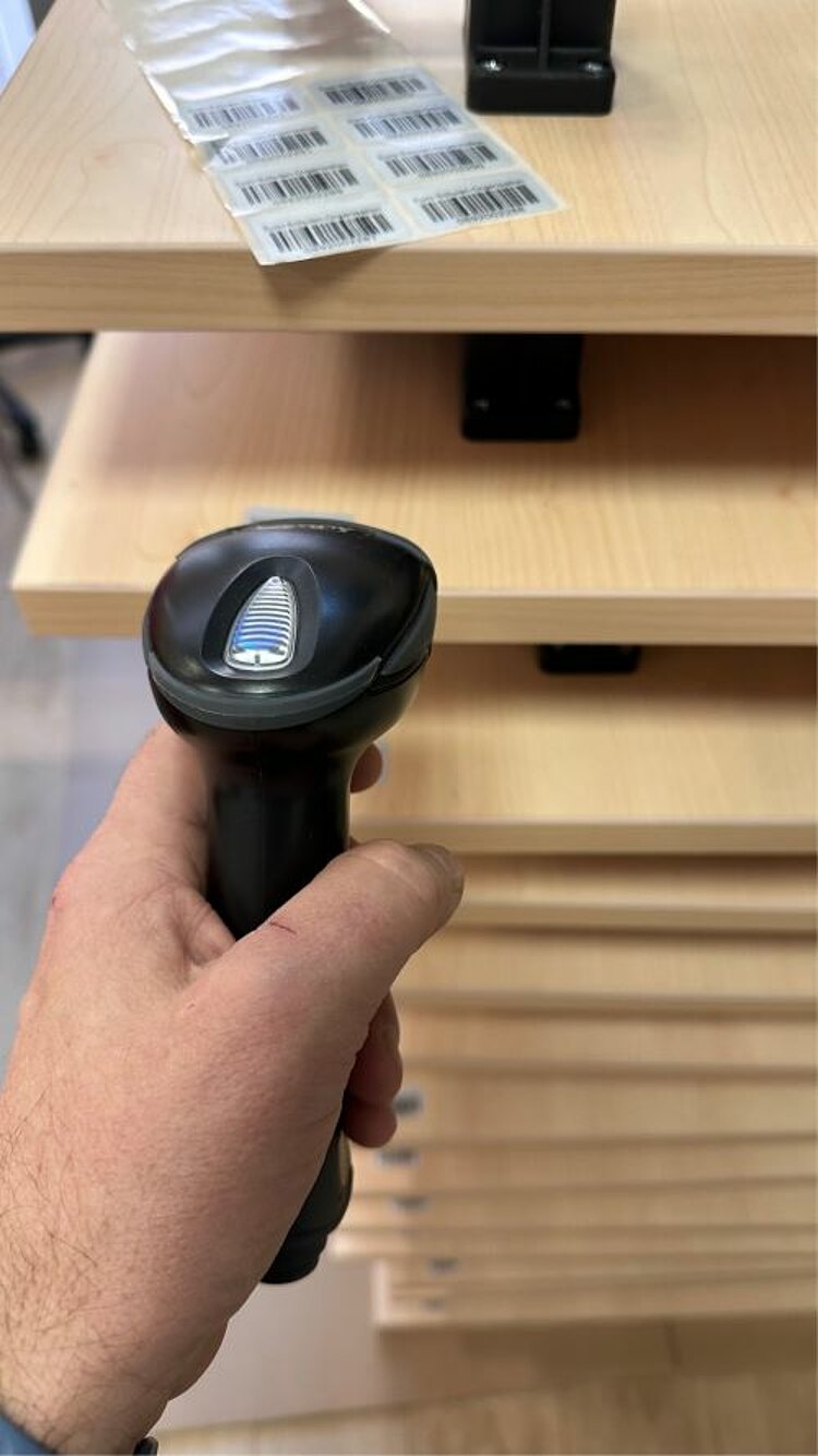 Frischer Wind im Klassenzimmer Ein Barcodescanner wird von einer Hand gehalten und ist auf mehrere gestapelte Tische aus Holz gerichtet. Auf der obersten Tischplatte liegt ein Blatt mit mehreren Barcodes.