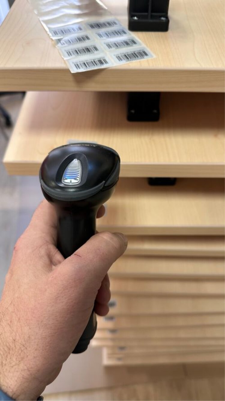 Ein Barcodescanner wird von einer Hand gehalten und ist auf mehrere gestapelte Tische aus Holz gerichtet. Auf der obersten Tischplatte liegt ein Blatt mit mehreren Barcodes.