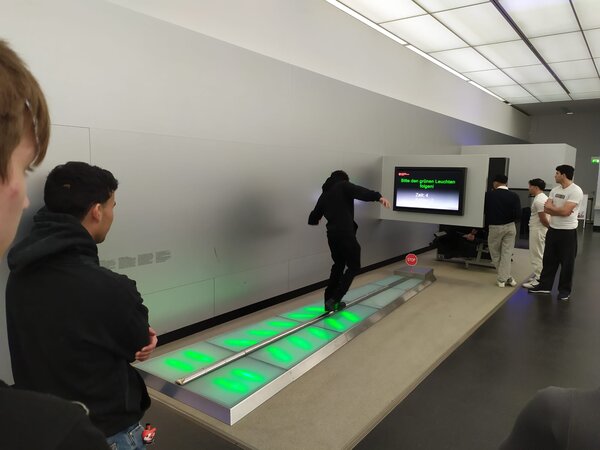 Eine Person testet ihre Reflexe auf einer beleuchteten Laufbahn in einem Technik- oder Wissenschaftsmuseum. Andere Personen beobachten das Experiment.