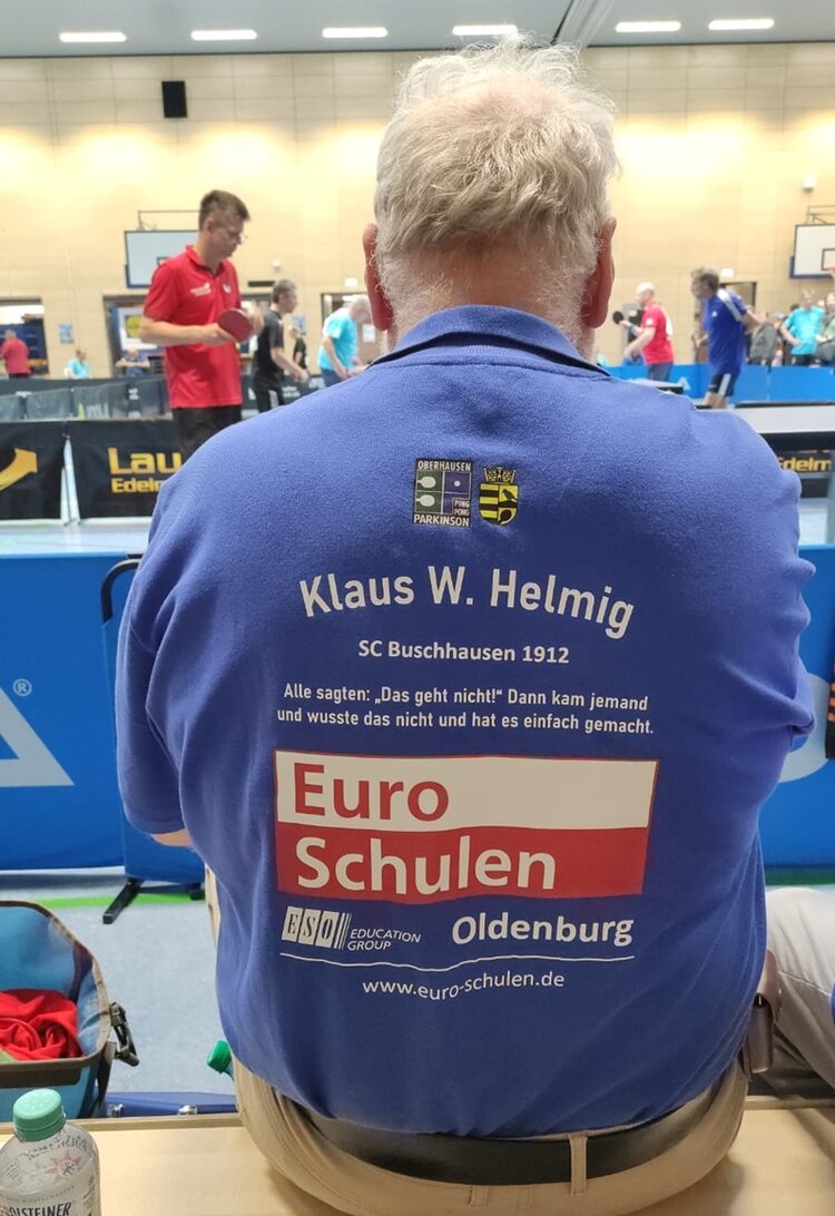 Ein männlicher Teilnehmer an den PingPongParkinson German Open der Euro-Schulen Oldenburg sitzt auf einer Bank, mit dem Rücken zur Kamera, der rot-weiße Euro-Schulen-Aufdruck auf blauem T-Shirts ist zu sehen.