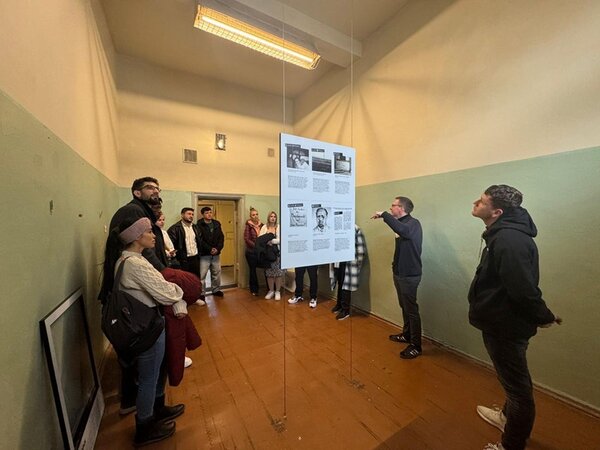 Die Gruppe der Teilnehmenden des Integrationskurses steht in einem schmalen Raum mit grün-beigen Wänden und braunem Holzboden. In der Mitte des Raumes hängt eine Infotafel mit mehreren Textabschnitten und Schwarz-weiß-Fotografien an transparenten Seilen von der Decke. Eine Person zeigt mit der Hand auf die Tafel, während die anderen aufmerksam zuhören. Die Beleuchtung erfolgt durch eine längliche Deckenleuchte. An der Wand lehnt ein gerahmtes Bild oder eine Tafel.