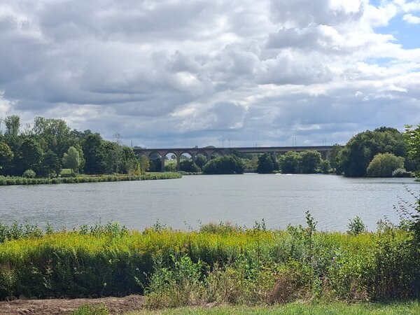 Weiter Blick über einen See mit üppiger Ufervegetation und einer großen Bogenbrücke im Hintergrund unter bewölktem Himmel.