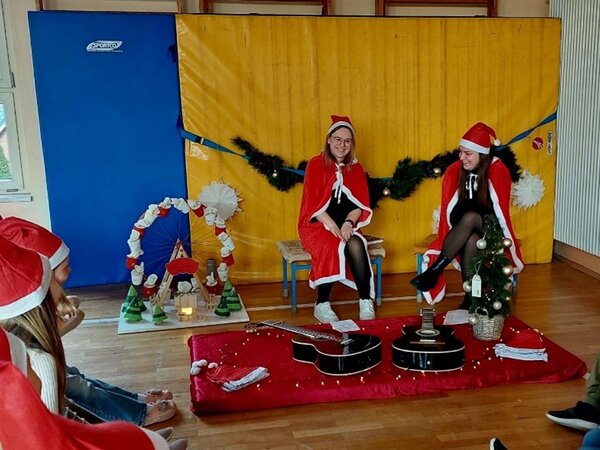Weihnachtlich geschmückter Bewegungsraum: Zwei als Weihnachtsfrauen verkleidete Studierende sitzen auf einer kleinen Bühne, vor ihnen Gitarren.