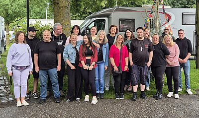 Das Gruppenfoto zeigt die Mitarbeitenden der Euro-Schulen Niederrhein auf dem Campingplatz Düsseldorf, wo sie sich unter einem Baum vor einem Campingmobil positioniert haben.