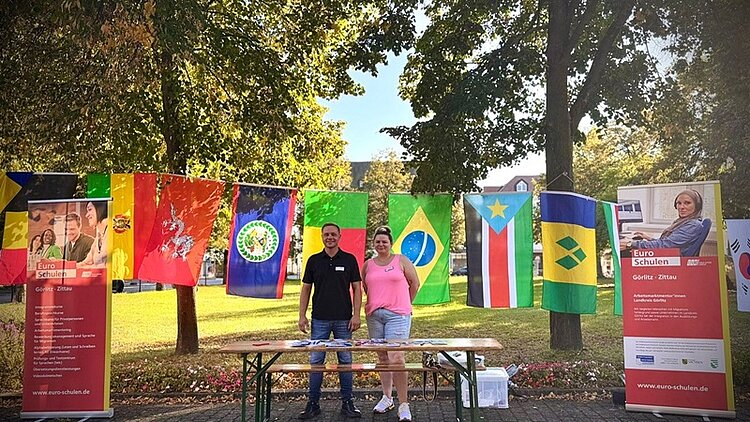 50 Jahre Interkulturelle Woche – 50 Jahre Einsatz für Vielfalt! Zwei Mitarbeitende der Euro-Schulen Görlitz posieren an ihrem Stand auf der Interkulturellen Woche. Links und rechts stehen große Euro-Schulen-Aufsteller, hinter ihnen ist eine Girlande aus internationalen Flaggen gespannt.