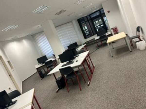 Büro mit mehreren Schreibtischen, Stühlen und Computern in einem hellen Raum mit Teppichboden und großen Fenstern mit vertikalen Lamellen.