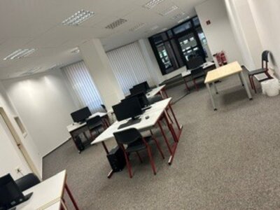 Büro mit mehreren Schreibtischen, Stühlen und Computern in einem hellen Raum mit Teppichboden und großen Fenstern mit vertikalen Lamellen.