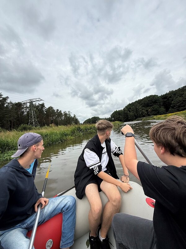 Drei junge Männer in einem roten Schlauchboot auf einem Fluss. Einer von ihnen paddelt, während der andere nach vorne schaut. Der Himmel ist bewölkt, und im Hintergrund sind Bäume und Sträucher zu erkennen.