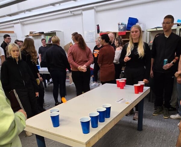 Mehrere Teilnehmende stehen um einen Tisch mit roten und blauen Bechern und spielen gemeinsam ein Geschicklichkeitsspiel.