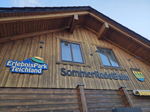 Holzgebäude mit der Aufschrift ‚ErlebnisPark Teichland‘ und ‚SommerRodelBahn‘, fotografiert von unten vor blauem Himmel.