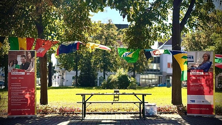 50 Jahre Interkulturelle Woche – 50 Jahre Einsatz für Vielfalt! Der Stand der Euro-Schulen Görlitz und Zittau auf der Interkulturellen Woche. Links und rechts stehen große Euro-Schulen-Aufsteller, in der Mitte steht ein Tisch, dahinter ist eine Girlande aus internationalen Flaggen gespannt. Die Sonne scheint, es ist eine grüne Wiese zu sehen sowie Bäume an den Seiten.