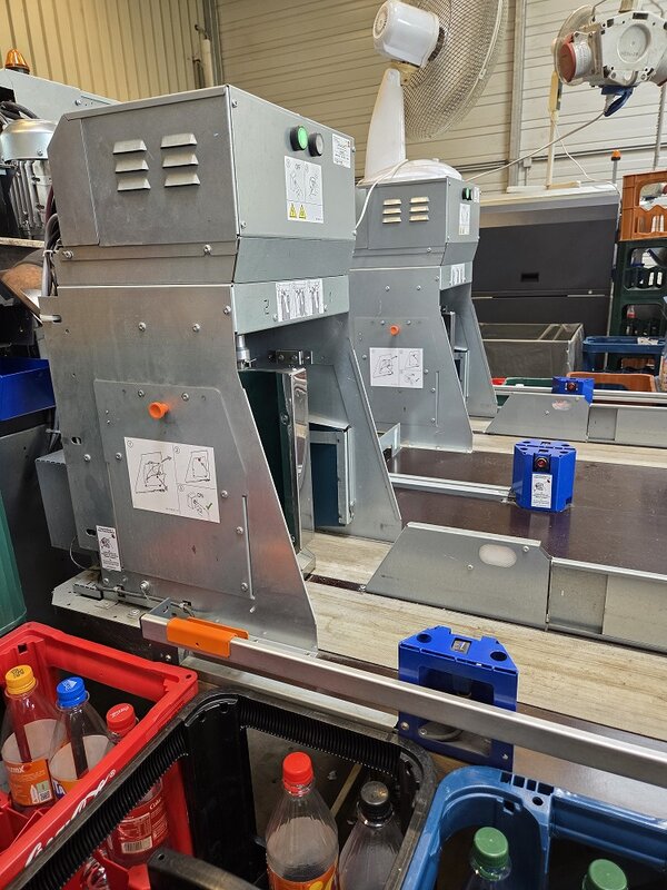 Industrielle Maschinen zur Verarbeitung und Verpackung von Lebensmitteln in einem Produktionsbetrieb; im Vordergrund stehen Förderbänder und Kunststoffkisten mit Getränkeflaschen.