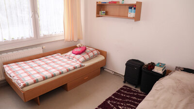 Kleines, schlichtes Einzelzimmer mit Holzbett, kariertem Bettzeug, Fenster mit Vorhängen, Wandregal und Gepäckstücken neben dem Bett.