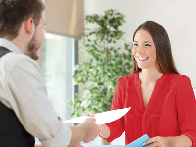 Eine Frau in einem roten Kleid sitzt an einem Tisch und lächelt, während sie einem Mann, der ihr gegenübersitzt, Dokumente übergibt. Der Mann trägt ein weißes Hemd und scheint die Papiere entgegenzunehmen. Die Szene deutet auf ein formelles oder berufliches Treffen hin.