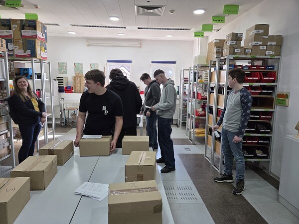 In einem hellen Raum mit Regalen voller Pakete steht in der Mitte ein Tisch mit mehreren Päckchen, drumherum stehen mehrere junge Menschen mit Zetteln in den Händen.
