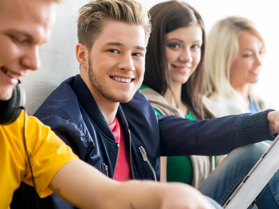 Ein lächelnder junger Mann mit einem blauen Jackett und einem rosa T-Shirt sitzt in einem Auto und hält das Lenkrad. Im Hintergrund sind drei weitere Personen, die ebenfalls lächeln und sich unterhalten.