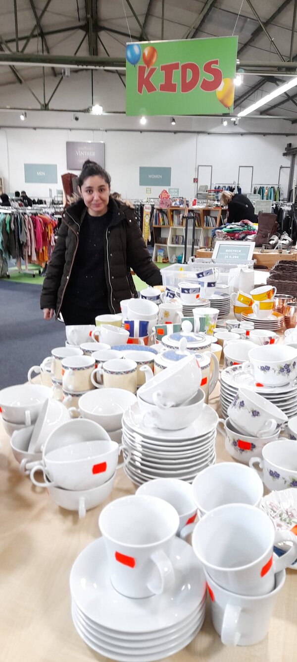 Eine Frau steht in einer Halle vor einem Tisch mit Geschirr und Tassen; über ihr hängt ein grünes Schild mit der Aufschrift „KIDS“.