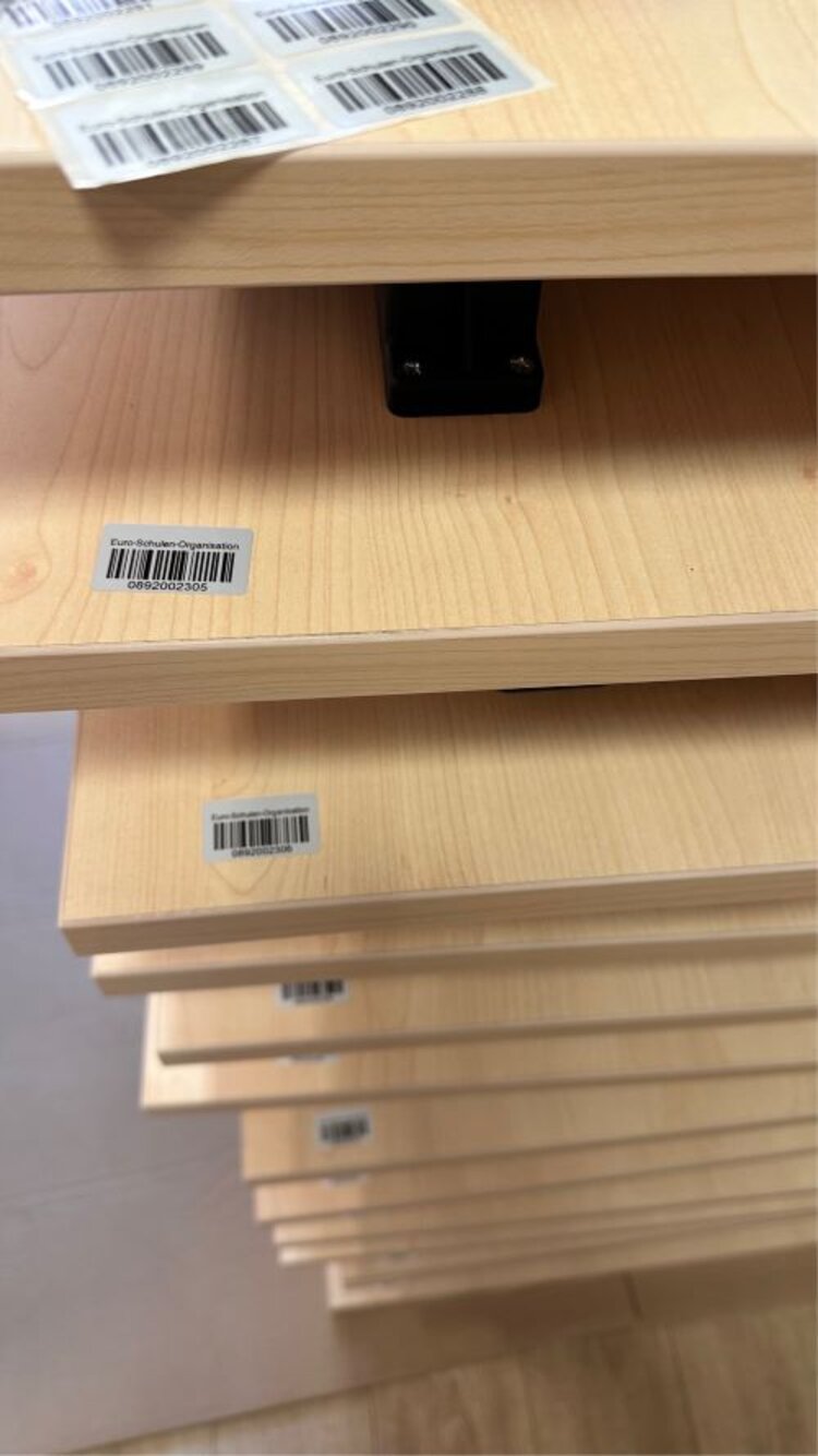 Mehrere Tische sind zusammengeklappt aufeinandergestapelt. Sie sind mit Barcodes versehen. Auf der obersten Tischplatte liegt ein Blatt mit mehreren Barcodes.