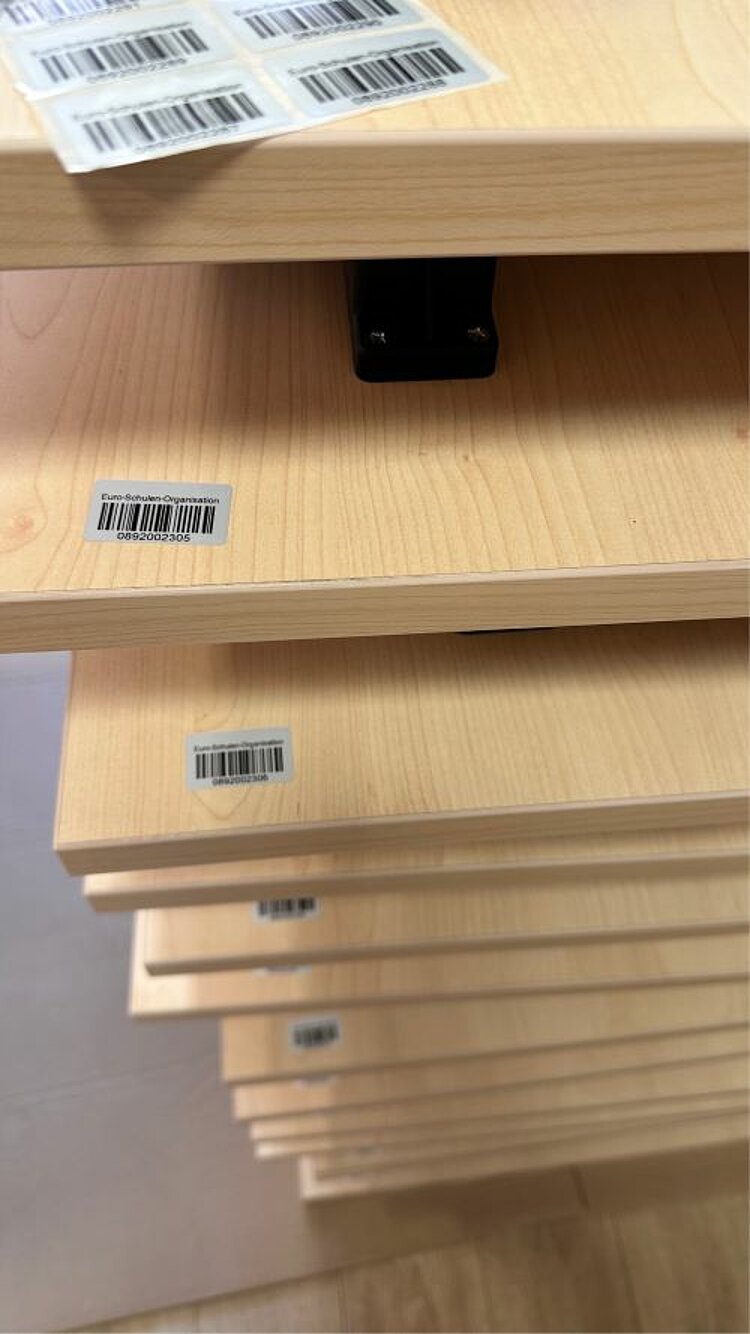 Frischer Wind im Klassenzimmer Mehrere Tische sind zusammengeklappt aufeinandergestapelt. Sie sind mit Barcodes versehen. Auf der obersten Tischplatte liegt ein Blatt mit mehreren Barcodes.