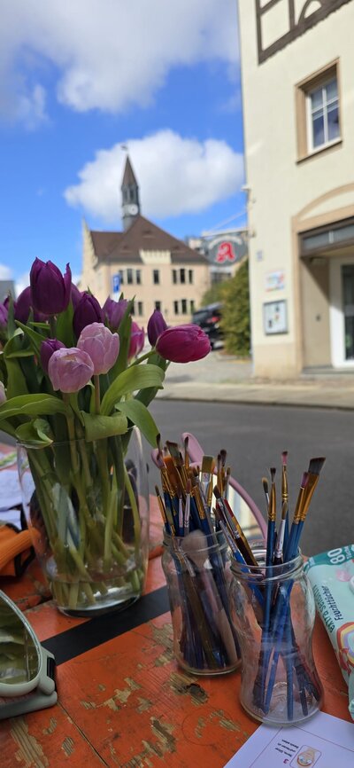 Auf einem Tisch stehen ein Strauß Tulpen sowie Gläser mit Pinseln; im Hintergrund ist eine Altstadtstraße zu sehen.