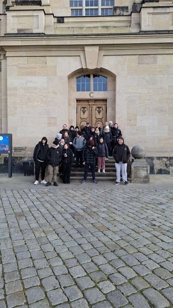 Eine Gruppe von Menschen steht für ein Gruppenfoto vor dem Eingang der Dresdner Frauenkirche, auf dem Vorplatz mit Kopfsteinpflaster