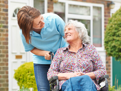 Eine Pflegekraft in blauer Uniform unterstützt eine ältere Frau im Rollstuhl, die ein buntes Blumenmusterhemd trägt. Beide wirken fröhlich und befinden sich im Freien vor einem Haus mit Ziegelmauer und grünen Pflanzen. Das Bild veranschaulicht die liebevolle und professionelle Betreuung im Rahmen der Qualifizierung zur Betreuungskraft § 53b (in Verbindung mit § 43b) SGB XI inklusive Demenzbegleitung.