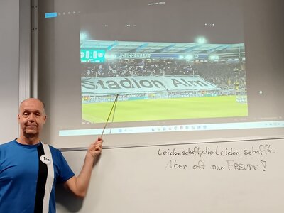 Dozent Ulrich im Arminia Bielefeld Trikot zeigt zum an die Wand projezierten Fußballspiel.