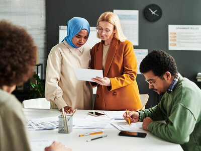 Drei Personen sind in einem Büro bei einer Besprechung zu sehen. Eine Frau mit einem blauen Kopftuch und eine ältere Frau mit Brille und orangefarbenem Blazer stehen nebeneinander und betrachten ein Dokument. Die ältere Frau hält ein Blatt Papier in der Hand. Ein junger Mann mit kurzem, lockigem Haar sitzt am Tisch und macht sich Notizen. Auf dem Tisch liegen verschiedene Unterlagen, Stifte, ein Smartphone und ein Becher mit Stiften. Im Hintergrund sind eine Pflanze, eine Uhr und ein Whiteboard zu erkennen.